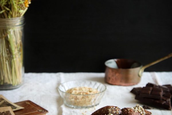 Plate of chocolate madeleines on a table with vintage photos, books, flowers, and dark chocolate pieces.
