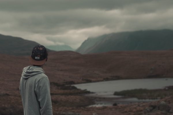 A person in a hoodie and cap looks at a mountain landscape under cloudy skies.