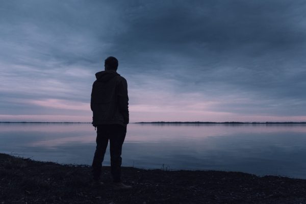 Silhouette of a person standing by a calm lake during a cloudy, pastel-colored sunset.