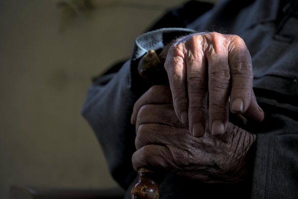Close-up of an elderly persons wrinkled hands resting on top of a wooden cane, with soft lighting.