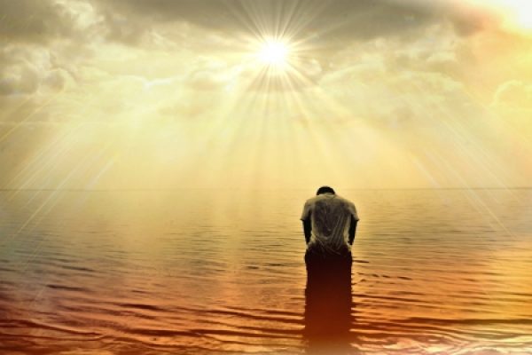 a person facing away and sitting in a body of water is bowing their head beneath a golden sky
