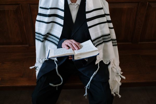 A person wearing a tallit reads from an open book while seated on a wooden bench.