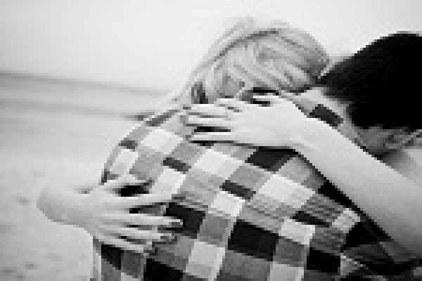 A couple embraces closely on a beach, with the ocean and sky in the background.