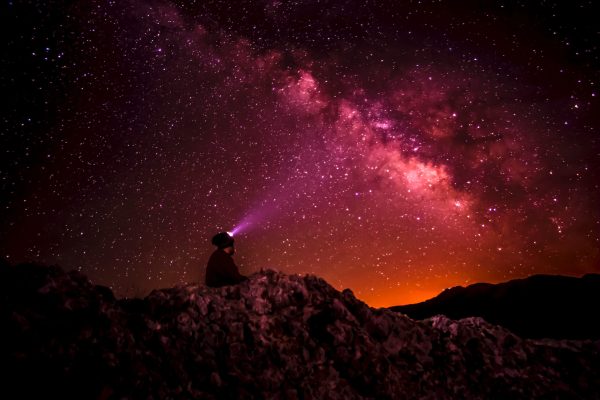 pinkish dark night sky with clouds and stars and person sitting on a boulder looking up at sky with headlight on their head