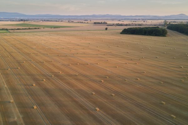 Aerial view of a vast harvested field with scattered hay bales, bordered by distant mountains under a clear sky.