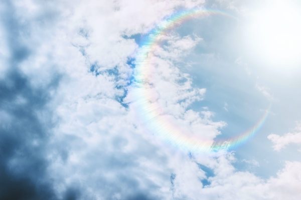 A rainbow halo surrounds the sun, partially obscured by clouds in a bright blue sky.