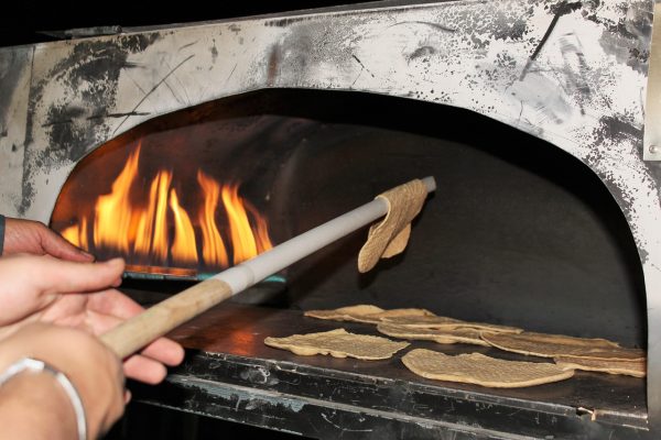 Hands place flatbread into a wood-fired oven using a long paddle, with flames visible inside.