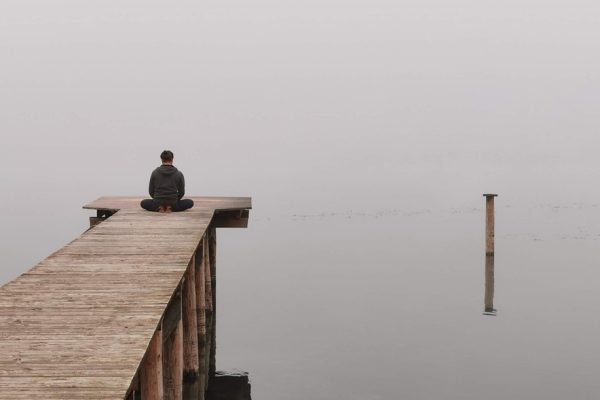 Person sitting on a wooden dock, facing a misty, calm lake.