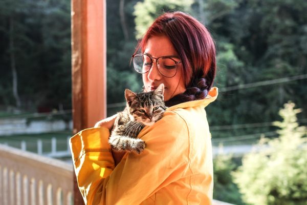 Woman with glasses and purple hair holds a tabby kitten on her shoulder outdoors, wearing a yellow jacket.