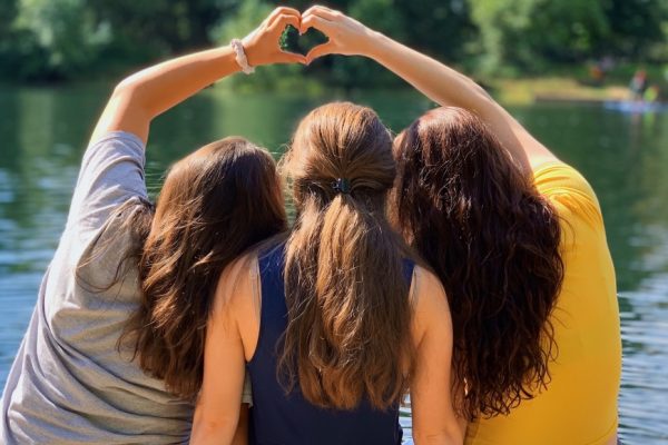 Three friends sit by a lake, forming a heart shape with their arms on a sunny day.