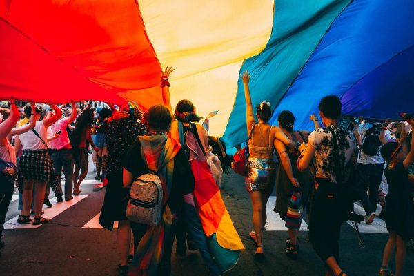 People walking under a large rainbow flag at a parade, celebrating LGBTQ+ pride and diversity.