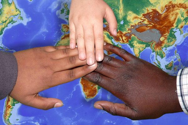 three hands--one with brown skin, one with white skin, one with black skin touch each other over the image of a world map