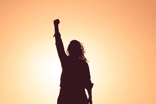 Silhouette of a person with curly hair raising a fist against a bright orange sunset background.