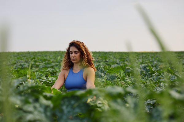 A woman with curly hair meditates in a lush green field under a clear sky.