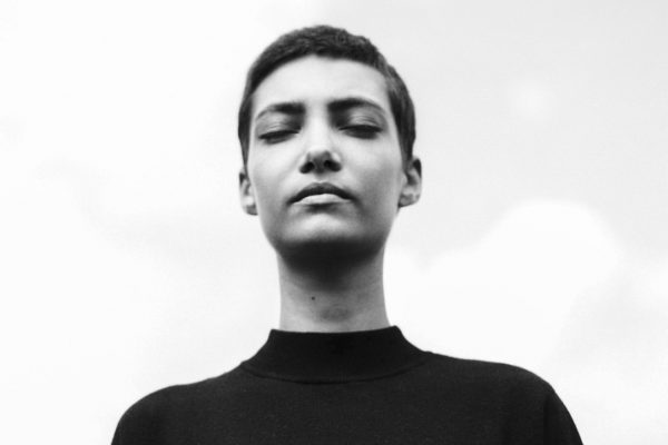 Black and white photo of a person with short hair, eyes closed, wearing a dark top, against a cloudy sky.