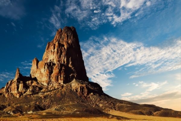 A towering rock formation under a blue sky with scattered clouds, set in a desert landscape.
