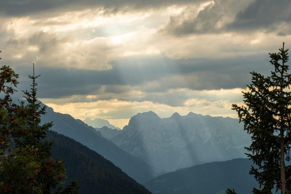 Mountain landscape with dramatic skies, sunbeams breaking through clouds, and silhouetted pine trees in the foreground.