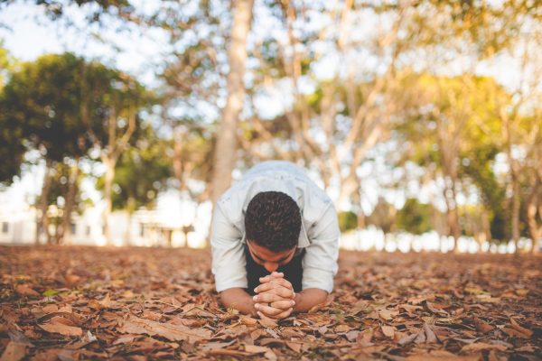 Person kneeling and praying on a bed of dry leaves in a park with trees in the background.