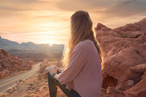 A woman sits on a rocky cliff, gazing at a sunset over a desert landscape with a winding road below.