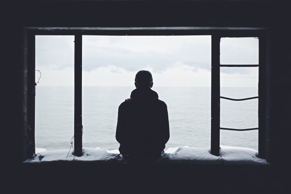 Silhouette of a person sitting in a window frame overlooking a calm sea under a cloudy sky.