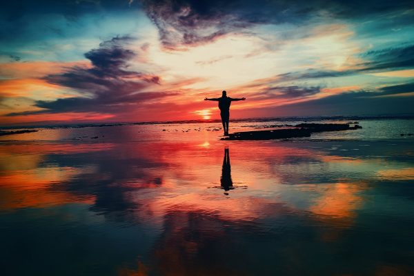 Silhouette of a person with arms outstretched against a vibrant sunset over a reflective body of water.