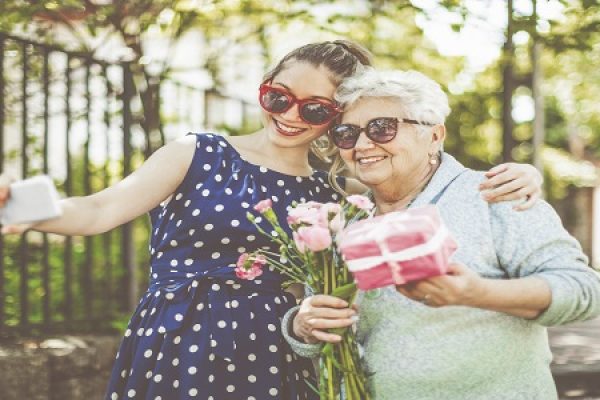 Two women wearing sunglasses, taking a selfie outdoors; one holds flowers, the other a gift.