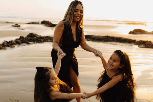 brown skinned woman in black tank dress holding hands and dancing with two brown skinned girls by the ocean with water in the background and sunlight