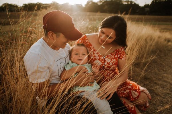 brown-skinned man and woman sitting in a wheat field with sun shining, holding cute toddler between them