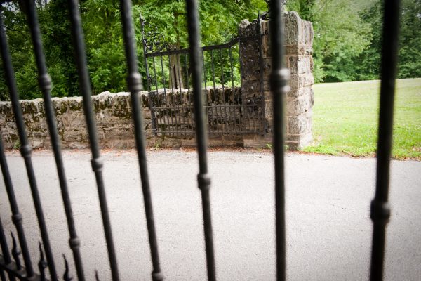 Wrought iron gate and stone wall viewed through black fence bars, with grass and trees in the background.