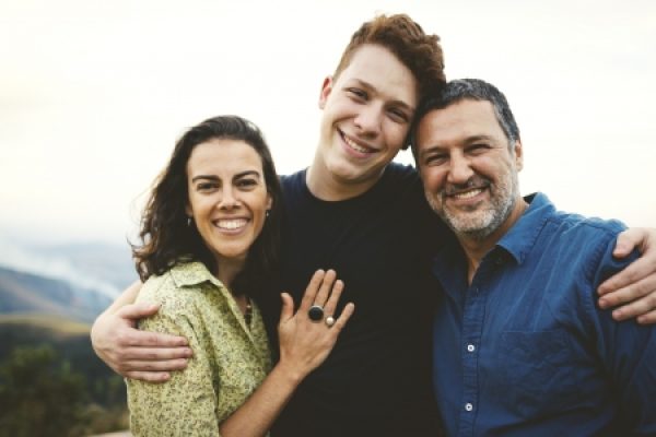 A family of three smiling together outdoors, with hills visible in the background.