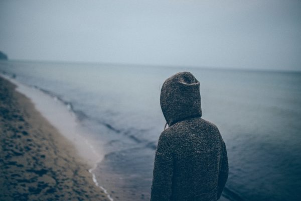 Person in a hoodie looking out at a calm, overcast seascape from a sandy beach.