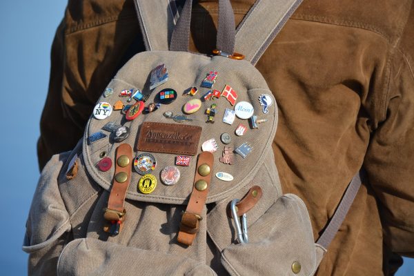 A brown backpack covered in various pins, worn by someone in a brown jacket.