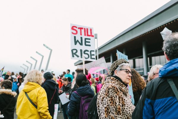 A protest crowd holding signs, including one that reads STILL WE RISE, outside a modern building.
