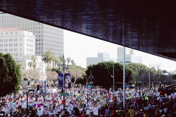 A large crowd gathers outdoors holding colorful protest signs in a city surrounded by trees and tall buildings.