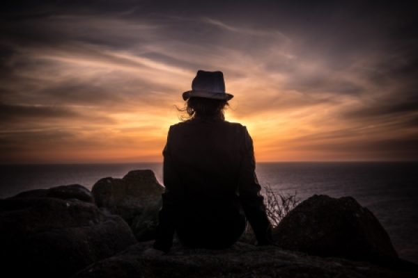 Silhouette of a person in a hat sitting on rocks, watching a dramatic sunset over the ocean.