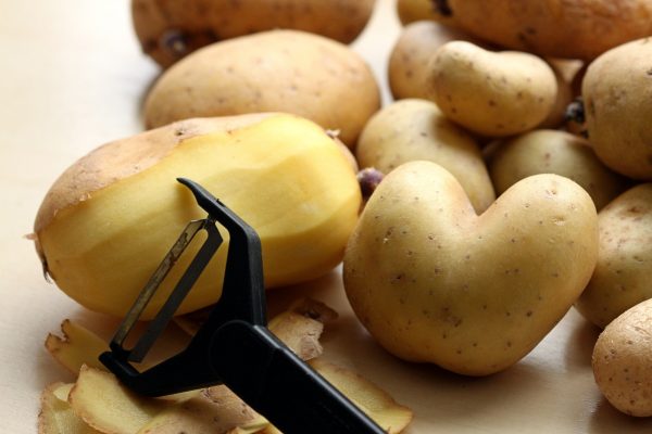 Potatoes with a peeler, one partially peeled, on a wooden surface. Some potatoes are heart-shaped.