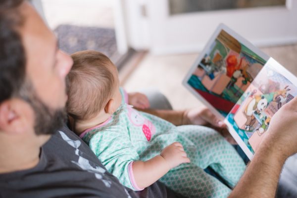 An adult reads a colorful book to a baby in pajamas on their lap.