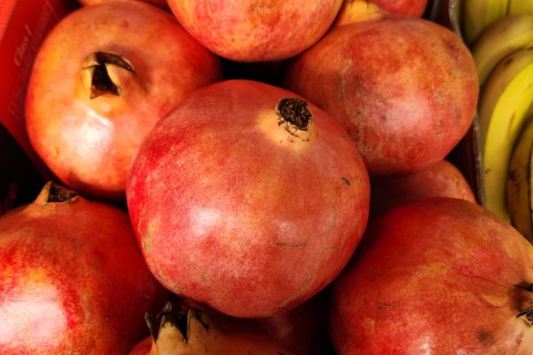 A pile of ripe pomegranates fills a box, with yellow bananas visible in the background.