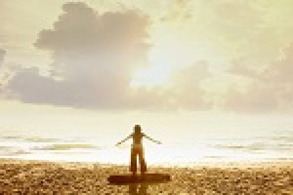 Person standing on a beach at sunset, arms outstretched, facing the ocean with a cloudy sky.