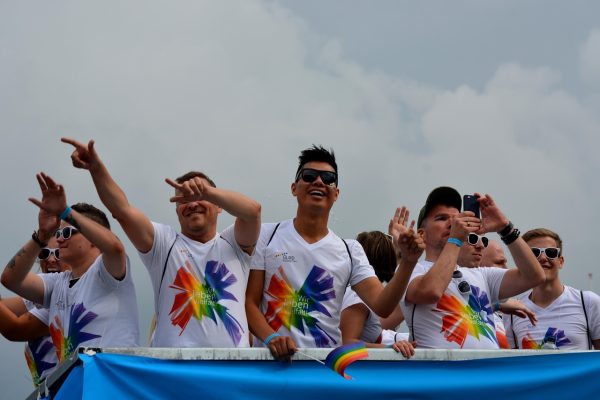 Group wearing rainbow shirts and sunglasses on a float at a parade, pointing and smiling under a cloudy sky.