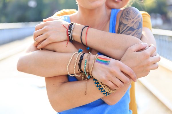Two people embracing, wearing colorful bracelets. One has a rainbow painted on their hand.