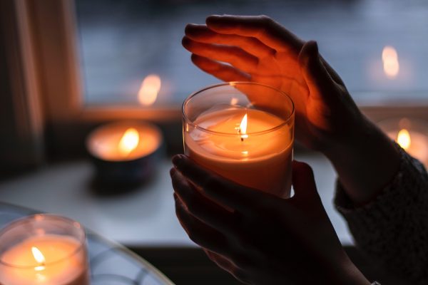 Hands cupping a lit candle in a glass jar, with more candles softly glowing in the background by a window.