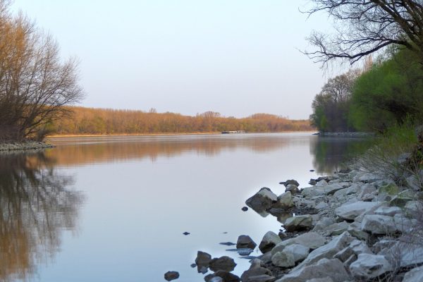 A serene river scene with still water, rocks in the foreground, and trees lining both banks under a clear sky.