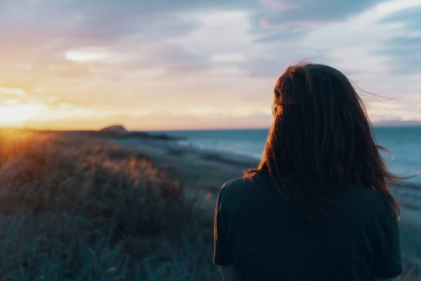 woman with long straight brown hair shown from behind looking toward body of water and shore of wheat and sky of clouds in pink and yellow and grayish.