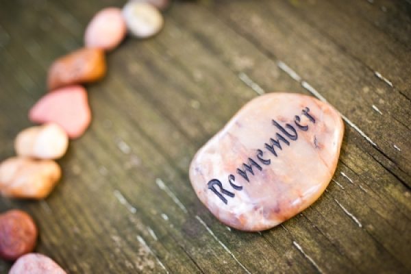 A smooth stone with Remember engraved on it lies on a wooden surface, surrounded by other colorful stones.