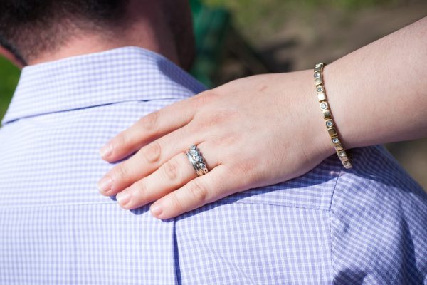 A hand with a ring and bracelet rests gently on a mans shoulder in a checkered shirt.
