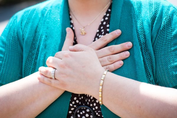 Person with hands on chest, wearing a green cardigan, polka dot top, gold bracelet, and necklace.