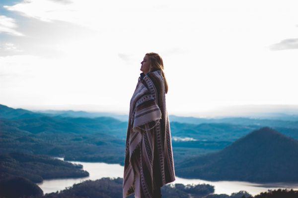 A person wrapped in a blanket stands overlooking a mountainous landscape with a lake and a cloudy sky.