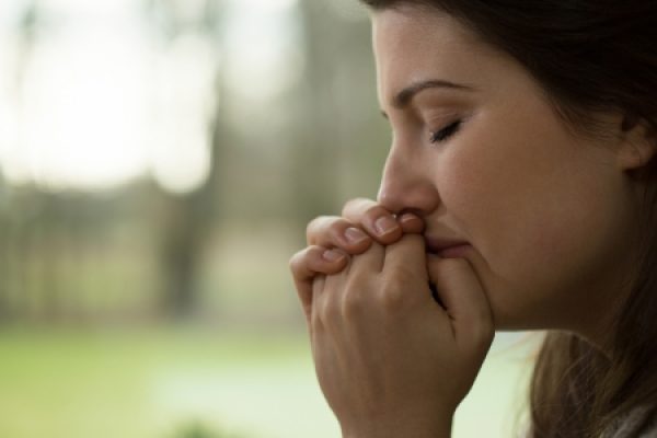 A woman with closed eyes and clasped hands in deep thought or prayer, with a blurred green background.