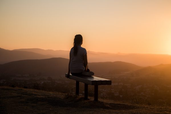 Person sitting on a bench overlooking hills at sunset, silhouetted against the sky.